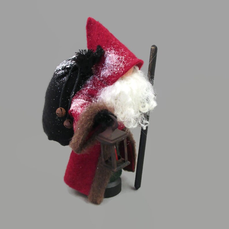 May include: A red felt Santa Claus doll with a white beard, black felt hat, and a black bag on his back. He is holding a lantern in one hand and a wooden stick in the other.