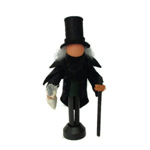 May include: A black felt doll with a top hat, white hair, and a black coat. The doll is holding a small white bag in one hand and a black stick with a brown tassel in the other hand. The doll is standing on a black base.