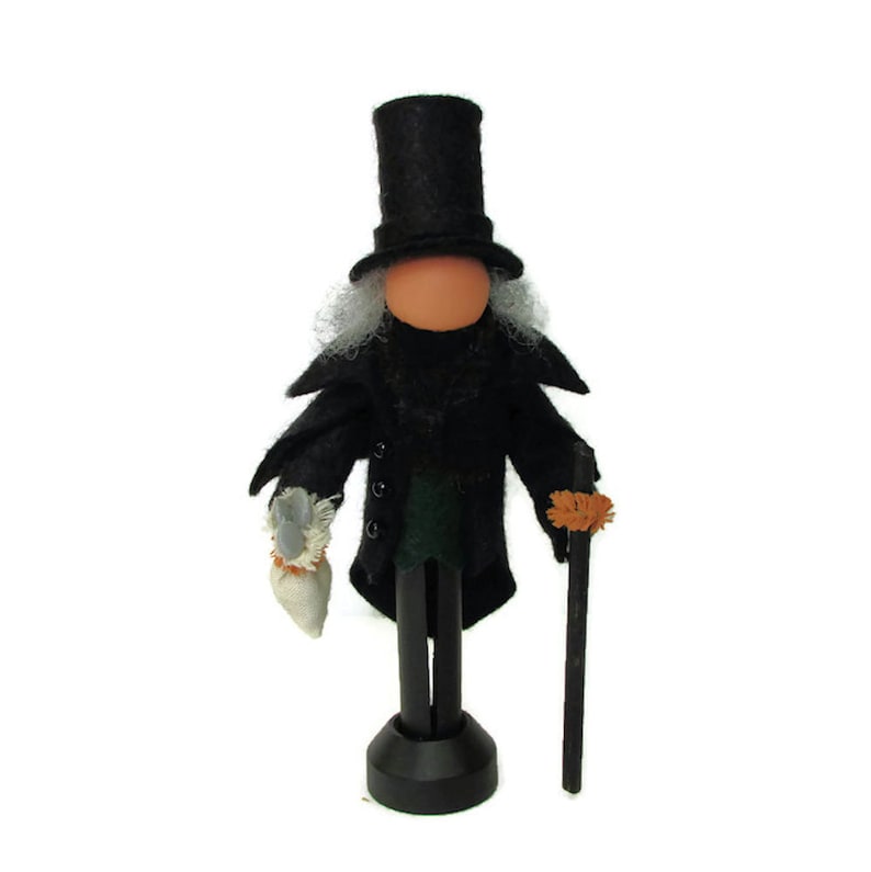 May include: A black felt doll with a top hat, white hair, and a black coat. The doll is holding a small white bag in one hand and a black stick with a brown tassel in the other hand. The doll is standing on a black base.