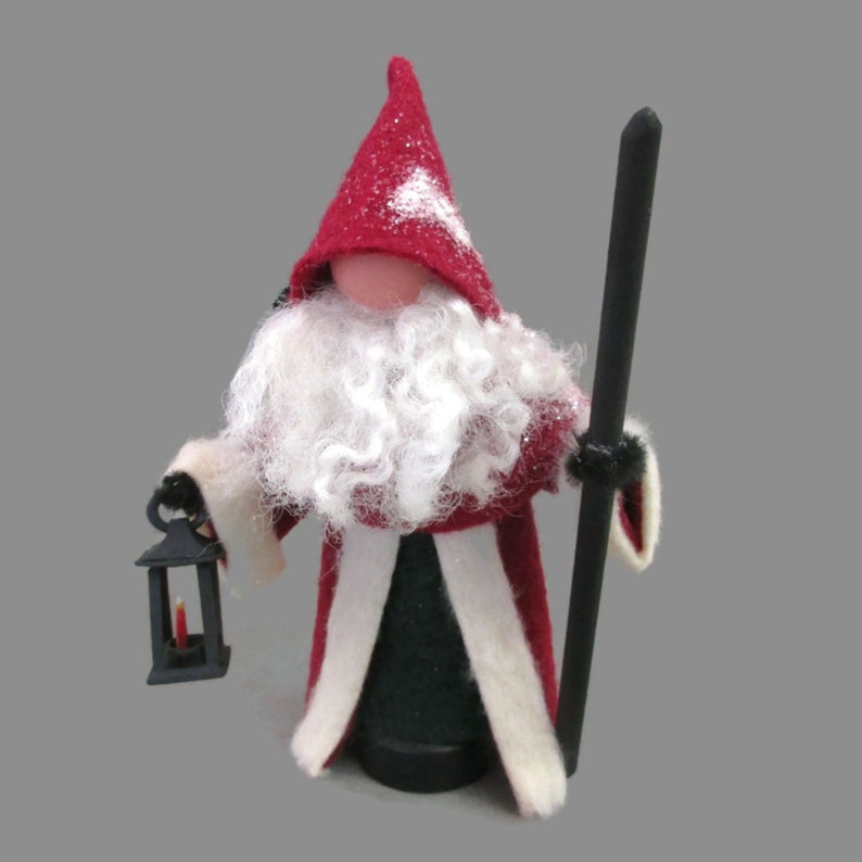 May include: A felt Christmas gnome wearing a red hooded robe with white trim, a white beard, and holding a black lantern and a wooden staff.