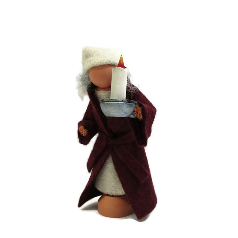 Pu&ograve; includere: Una bambola di feltro con un cappello bianco e un abito color borgogna, che tiene una piccola candela in un portacandele argentato.
