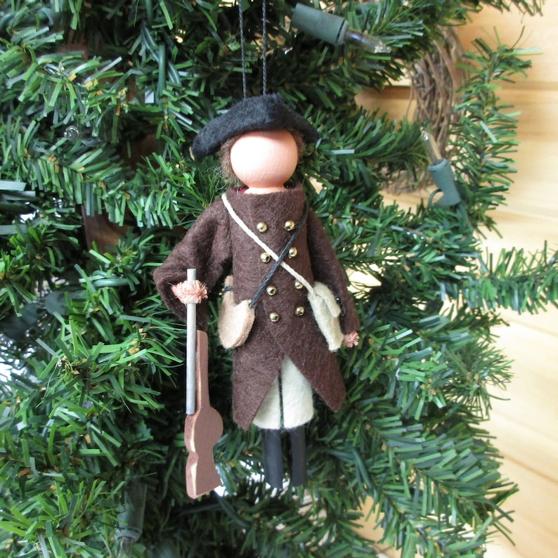 May include: A felt Christmas ornament depicting a person in a brown coat and black hat holding a brown rifle. The person has a white face and gold buttons on their coat.