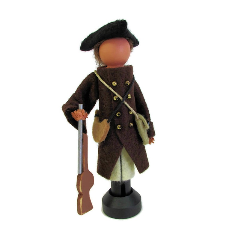 May include: A brown felt doll wearing a black hat and brown coat with gold buttons. The doll is holding a wooden gun and has a white face.