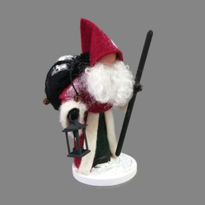 May include: A felt Christmas gnome figurine with a red hat, white beard, and black bag. The gnome is holding a black walking stick and a black lantern.