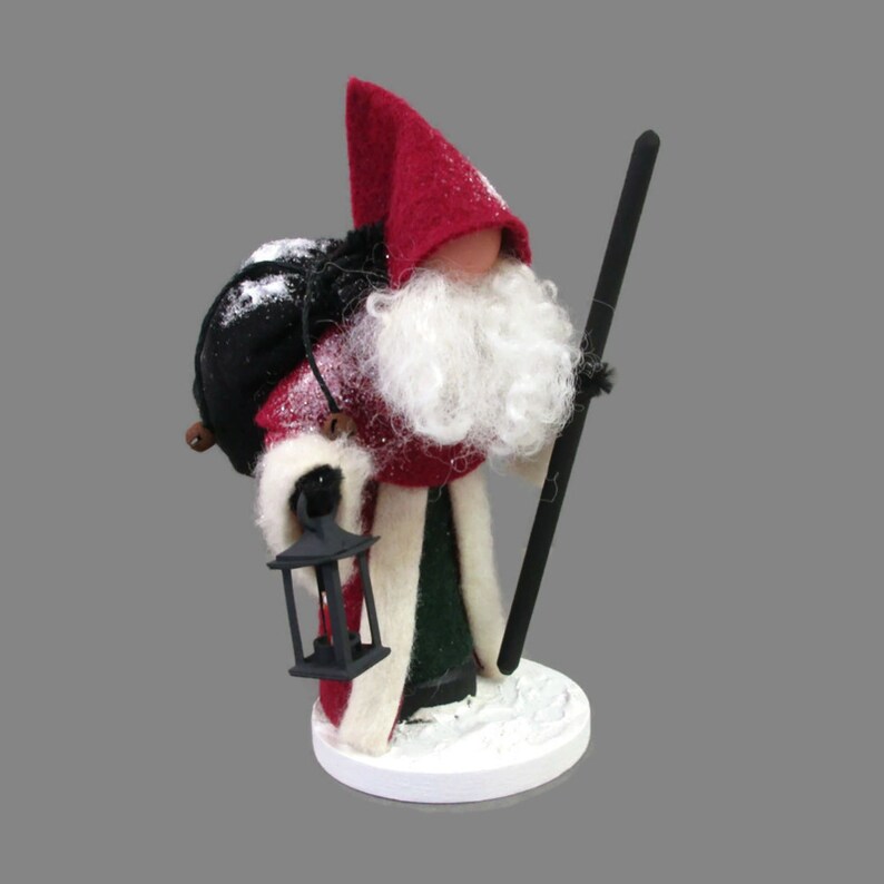 May include: A felt Christmas gnome figurine with a red hat, white beard, and black bag. The gnome is holding a black walking stick and a black lantern.
