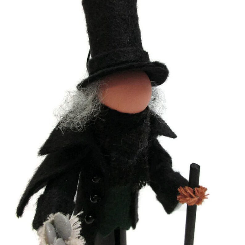 May include: A felt doll wearing a black top hat, black coat, and green vest. The doll has a white beard and is holding a black stick with a brown brush on the end.