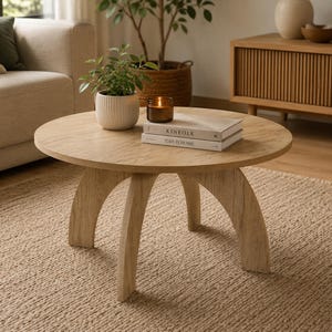 Puede incluir: Una mesa de centro redonda de madera clara con tres patas curvas. Sobre la mesa hay una maceta de cerámica blanca acanalada con una planta verde, un pequeño frasco de vidrio ámbar con una vela encendida y dos libros apilados titulados KINFOLK y THIS IS HOME.