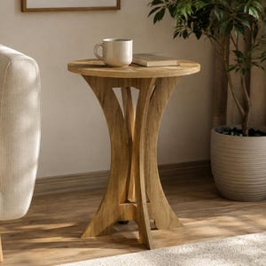 May include: A round wooden side table with a light brown finish, featuring a pedestal base with three curved legs that meet in the center. The table surface holds a beige ceramic mug and a small closed book, showcasing a smooth, natural wood grain.
