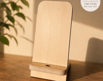 May include: A light-colored wooden phone stand with a smooth, vertical back panel and a horizontal base ledge featuring a small circular cutout. The image includes text that reads DIGITAL DOWNLOAD CNC FILES DXF SVG PDF and WOODEN PHONE STAND.
