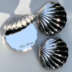 May include: Three silver-toned metal compact mirrors shaped like seashells. One mirror is open, revealing a reflective surface with the engraved text Mrs. Stevens, while the other two are closed, showing the ribbed, scalloped exterior shell design.