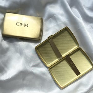 May include: Two rectangular gold-toned metal cases, one closed and one open. The closed case features the letters C&M engraved on the front. The open case reveals a textured interior with a central metal bar and a hinged clip for securing contents.