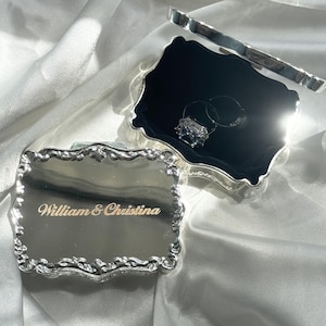 May include: A rectangular silver-toned metal box with an ornate, scalloped edge and a hinged lid. The lid is engraved with the text William & Christina. The interior is lined with black velvet and contains a silver ring with a clear stone.