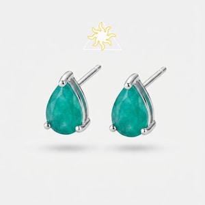 May include: A pair of silver stud earrings featuring pear-shaped, faceted green gemstones held by three prongs. Above the earrings is a small yellow line-art icon of a sun inside a triangle. The earrings have a post back for secure attachment.