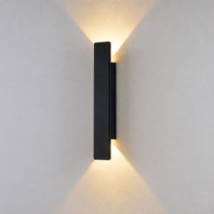Könnte beinhalten: Eine vertikale, rechteckige Wandleuchte mit mattschwarzer Oberfläche. Die Leuchte verfügt über eine integrierte Lichtquelle, die einen warmen, hellen Schein nach oben und unten wirft und zwei markante dreieckige Lichtkegel an der Wand erzeugt.