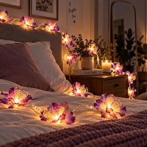 Könnte beinhalten: Eine Lichterkette mit dekorativen LED-Leuchten in Form von violetten und weißen Blüten. Jede Blüte enthält in der Mitte eine warm leuchtende Glühbirne, wobei dünne, lichtemittierende Filamente nach außen ragen und einen hellen Effekt erzeugen.