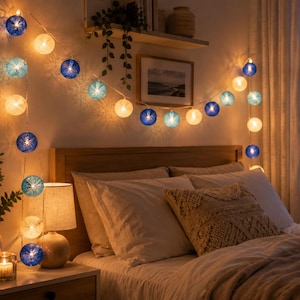 Peut inclure: Une guirlande lumineuse décorative composée d'abat-jours sphériques dans des tons bleu foncé, bleu clair et blanc chaud. Les lumières sont allumées, diffusant une lueur douce, et sont disposées en guirlande sur le mur au-dessus d'un lit.