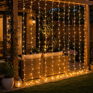 May include: A curtain of warm white LED string lights hanging vertically from a wooden pergola structure. The lights are arranged in multiple parallel strands, creating a glowing backdrop behind a wicker outdoor sofa with light cushions.
