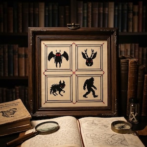 May include: A framed cross-stitch sampler featuring four black pixelated cryptid silhouettes with red eyes: a moth-like creature, a jackalope, a canine-like beast, and a bipedal ape-like figure, all arranged in a grid within a dark wooden frame.