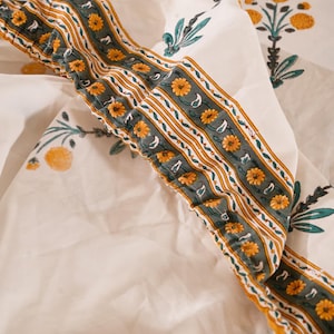 May include: A white fabric sheet featuring a repeating pattern of yellow flowers and green stems. The edge is finished with a wide border containing horizontal stripes of yellow, white, and green, accented with small yellow floral motifs.