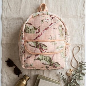 May include: A quilted backpack in a soft peach hue featuring a repeating pattern of green birds and white flowers on branches. The bag includes a top handle, a front pocket with a flap, and orange and white striped trim along the edges.