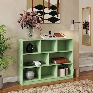 May include: A light green wooden bookcase with three horizontal shelves divided into eight compartments of varying sizes. The unit holds decorative items including a black sculpture, a dark tray with white gloves, a brown book, and a round box.