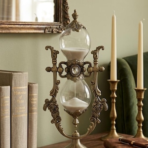 Puede incluir: Un reloj de arena de estilo antiguo con un marco de bronce metálico, tallas florales intrincadas y un cristal transparente central. El reloj contiene arena blanca en dos bulbos de vidrio y se apoya sobre una base circular con detalles de rosas.