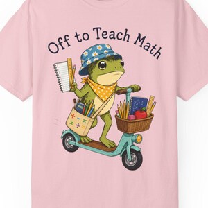 May include: A light pink t-shirt featuring a graphic of a green frog wearing a blue daisy-patterned bucket hat and a yellow polka-dot bandana. The frog rides a teal scooter, carrying a bag and basket filled with school supplies and the text Off to Teach Math.