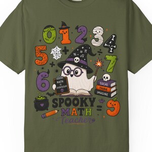 May include: An olive green t-shirt featuring a graphic of a ghost wearing a witch hat and glasses, holding a clipboard that says "MATH IS SPECTACULAR". The design includes numbers zero through nine, a stack of books, and the text "SPOOKY MATH TEACHER".