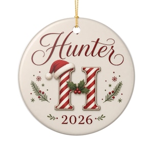 Puede incluir: Un adorno de cerámica redondo de color crema con el nombre Hunter en letra roja, una letra H grande con rayas rojas y blancas, un gorro de Papá Noel rojo, hojas de acebo verdes con bayas rojas y el año 2026 en rojo, con un cordón dorado.