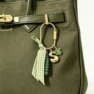 Puede incluir: Un bolso de mano de ante verde oliva con un llavero ovalado de metal dorado. El llavero está adornado con una cinta de cuadros vichy verde y blanca, una hilera de cuentas verde oscuro, un dije con la letra S dorada y un trébol de cuatro hojas.