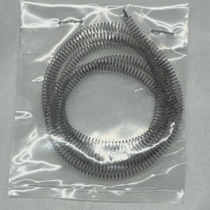 May include: A coiled metal heating element, gray in color, is enclosed in a clear plastic bag. The element is tightly wound in a spiral shape.