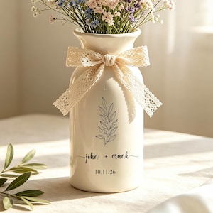 Può includere: Un vaso in ceramica color crema con un disegno a linee nere di un ramo, il testo "john + crank" e la data "10.11.26". Il vaso è decorato con un nastro di pizzo beige ed è riempito con un bouquet di margherite bianche e fiori di lavanda.
