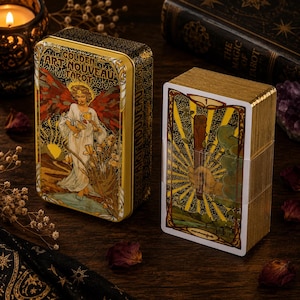 May include: A Golden Art Nouveau Tarot deck featuring a rectangular metal storage tin with an illustration of an angel holding two golden cups, and a stack of tarot cards wrapped in clear plastic showing a hand holding a wooden staff on the front.