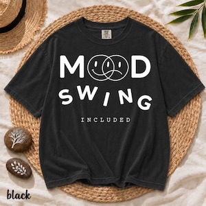 Puede incluir: Una camiseta negra de manga corta con texto blanco que dice MOOD SWING INCLUDED. El diseño incluye dos caras circulares superpuestas, una con expresión sonriente y otra con expresión triste, situadas entre las letras M y D.
