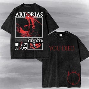 May include: A black acid-wash t-shirt featuring a graphic print. The back displays the text ARTORIAS, The Abyss Walker, and Japanese characters with a red illustration of a knight. The front features the red text YOU DIED and a red circular symbol.