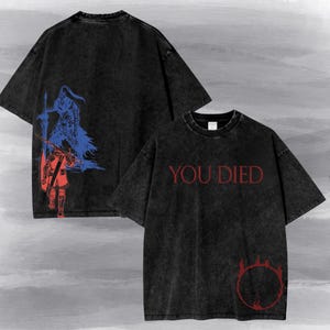 May include: A black acid-wash t-shirt featuring a front graphic with the red text "YOU DIED" above a red circular flame emblem. The back displays a blue and red illustration of two armoured figures with swords, one standing and one walking away.