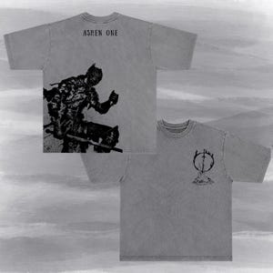 May include: A grey short-sleeve t-shirt featuring a black graphic of a knight holding a sword on the back with the text ASHEN ONE above it. The front of the shirt displays a smaller black graphic of a sword inside a circle above a pile of flames.