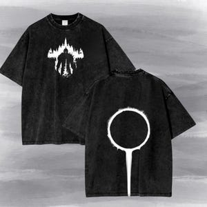 May include: A black acid-wash t-shirt featuring a white graphic on the front of a knight silhouette standing before a forest and castle. The back of the shirt displays a large white circular ring graphic with a vertical line extending downward from the base.
