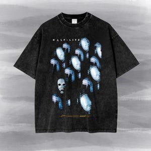 May include: A black acid-wash t-shirt featuring the text HALF-LIFE at the top and a graphic print of multiple blue-tinted gas mask figures. The design includes small logos at the bottom, including the Valve and Source engine branding.