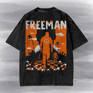 Può includere: Una t-shirt nera con effetto lavaggio acido e una grande grafica arancione sul retro. Il design mostra la scritta FREEMAN sopra la sagoma di una figura che tiene in mano un piede di porco, circondata da creature aliene e alti treppiedi.