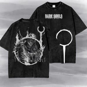 May include: A black acid-wash t-shirt featuring the text "DARK SOULS" in a dripping font on the front. The back displays a detailed white graphic of a knight in armour holding a sword, framed by a circular emblem and flame-like patterns.