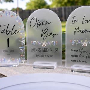 May include: Three arched frosted acrylic signs with clear rectangular bases. The first reads Table 1, the second Open Bar DRINKS ARE ON US! HANGOVER IS ON YOU!, and the third In Loving Memory We know You would be here if Heaven wasn't so far away.