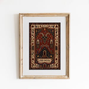 May include: A framed print of The Devil tarot card featuring a central goat head, broken chains, and two figures standing before a dark archway. The artwork is rendered in deep red, black, and cream tones with intricate border details and text.