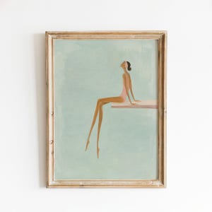May include: A framed art print featuring a stylized illustration of a person with dark hair wearing a pink swimsuit and headband, sitting on a diving board against a light blue background. The frame is made of light-colored wood with a simple profile.