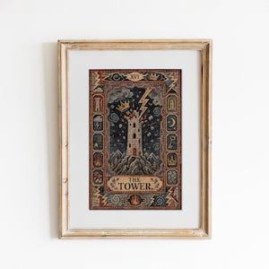 Op de afbeelding: Een ingelijste print van de tarotkaart De Toren met een stenen toren die door de bliksem wordt getroffen op een bergtop. Het ontwerp bevat een donkerblauwe lucht, een gouden kroon en een decoratieve rand met symbolen zoals sleutels, vlammen en wolken. De tekst luidt THE TOWER en XVI.