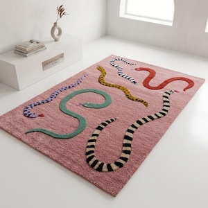 Puede incluir: Una alfombra rectangular de color rosa con un patrón de seis serpientes de varios colores y texturas, incluyendo morado, verde, amarillo, rayas negras y blancas, y rojo sólido, cada una con una pequeña lengua en la cabeza.