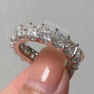 Könnte beinhalten: Ein silberfarbener Eternity-Ring mit einer durchgehenden Reihe klarer, quadratisch geschliffener Steine in einer Metallfassung. Der Schriftzug Silverfacetco ist auf der Metalloberfläche eingraviert und zeigt ein reflektierendes Finish.