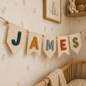 May include: A decorative banner featuring five off-white fabric pennants suspended on a string with wooden beads. Each pennant displays a single raised, textured letter in dark teal, mustard yellow, dusty blue, burnt orange, and sage green.