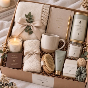 May include: A self-care gift box containing a cream-colored knit throw with a ribbon, a journal with a gold pen, a ceramic mug, a candle, herbal tea, bath salts, ritual chocolate, a wooden brush, hand cream, soap, and a green scrunchie.
