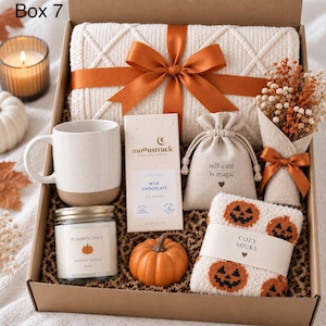 Könnte beinhalten: Eine Geschenkbox mit einer cremefarbenen Strickdecke, die mit einem orangefarbenen Band gebunden ist, einer weißen Keramiktasse, einer Moonstruck-Milchschokoladentafel, einem kleinen Beutel, einem Trockenblumenstrauß, einer Kürbiskerze, einem Kürbis und Socken.